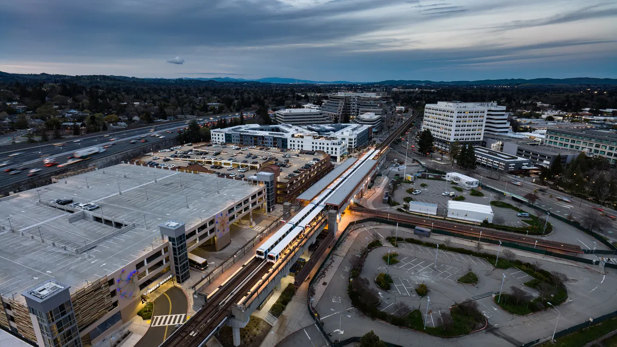 An aerial view of a train passing through a station next to a vacant parking lot under a cloudy sky.