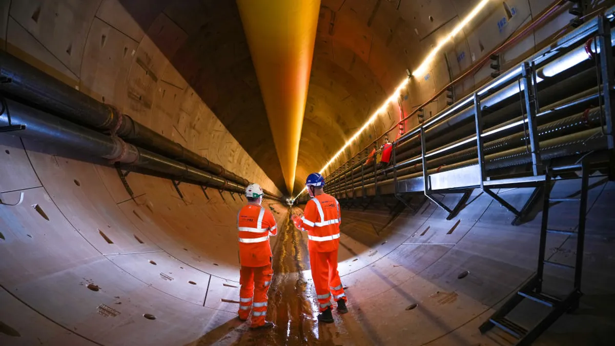 Two workers in orange suits with their backs to the camera stand in an industrial tunnel and observe its progress.