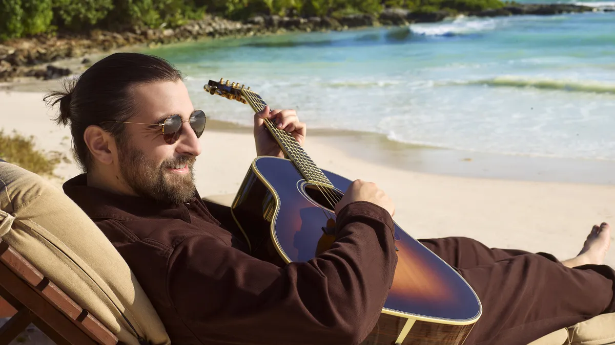 Singer Noah Kahan plays a guitar on the beach.