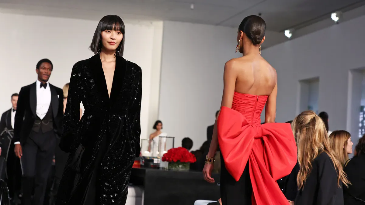 Models in red and black clothing pass each other on a runway during a fashion show as an audience watches.