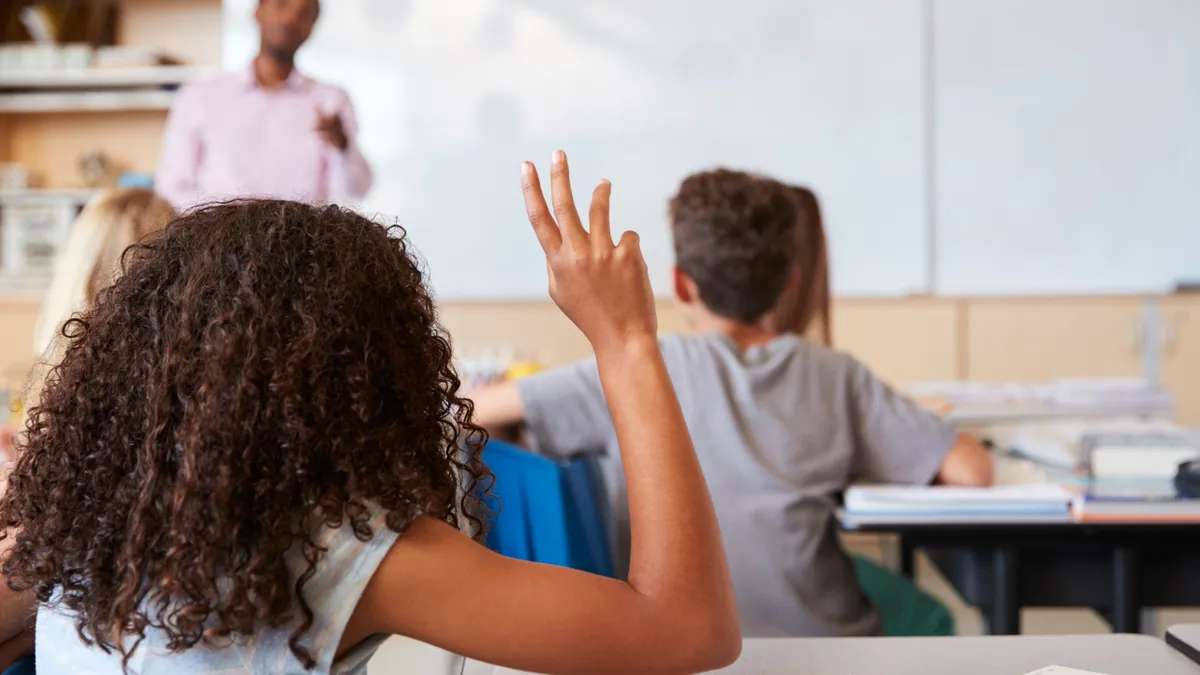 A student with back toward camera is sitting at a desk in a classroom and has an arm raised. In the background is another student and an adult standing at the front of the classroom.