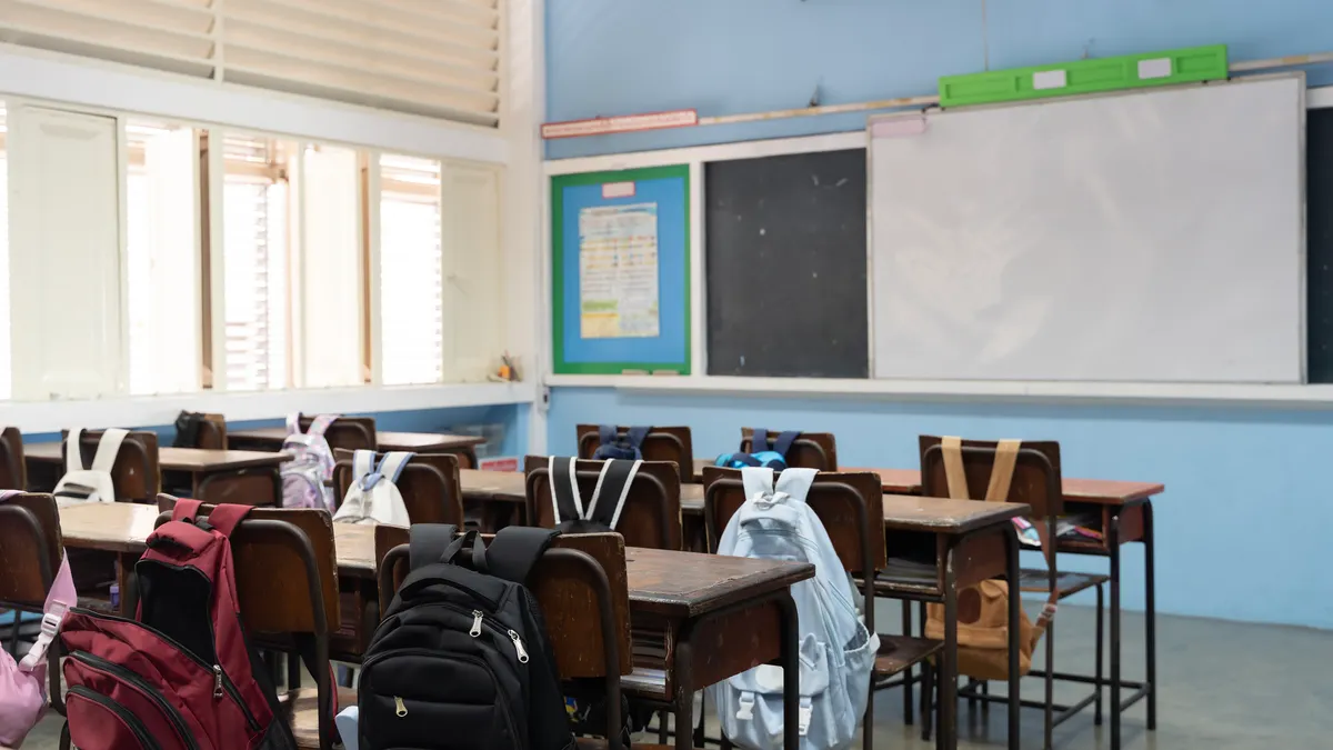 An empty classroom has backpacks rests on the back of chairs near desks.