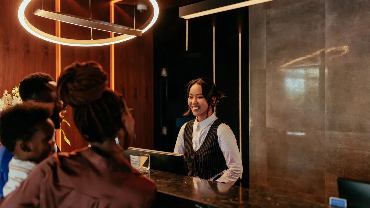 A young Asian receptionist is at the front desk of a luxurious hotel talking to her African American hotel guests.