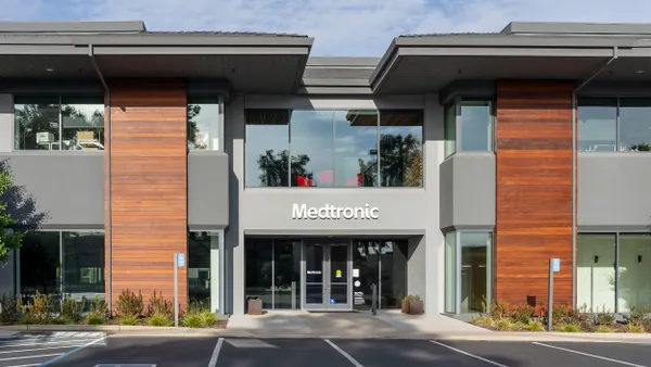 A modern two-story commercial office building with a mix of gray and wood paneling on the exterior. Large glass windows dominate both floors, and the front entrance features the "Medtronic" logo above double glass doors. The surrounding area includes a few small shrubs and empty parking spaces in front.