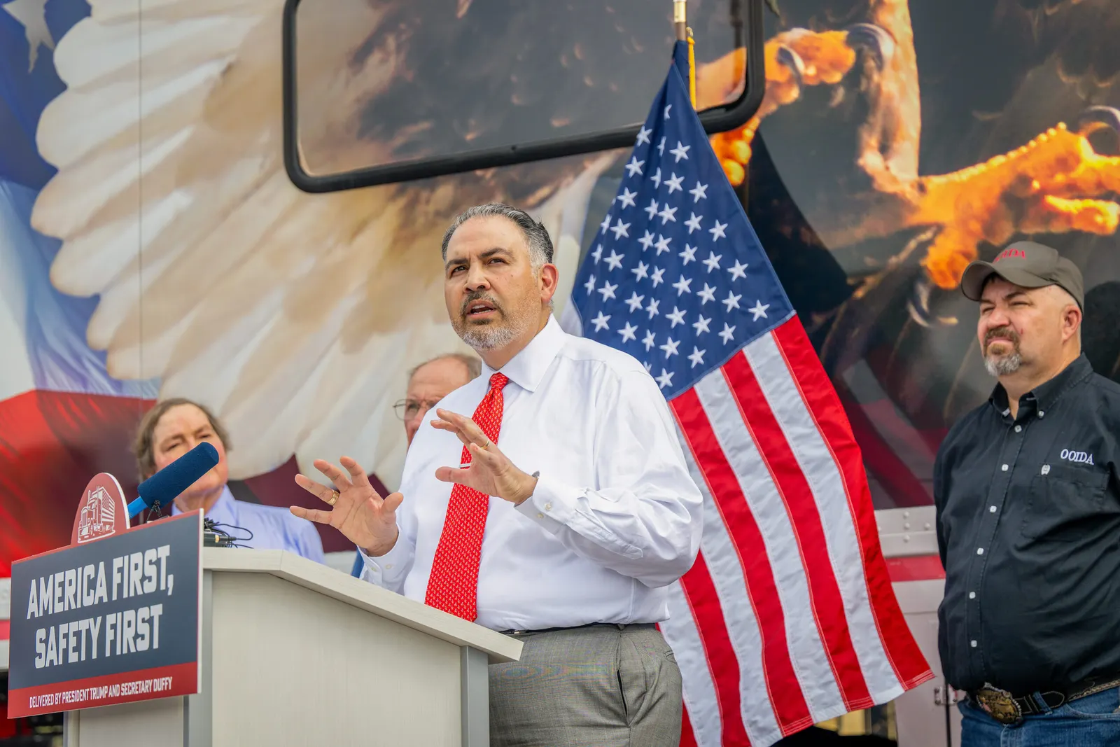 A person stands at a pulpit surrounded by three people and an American flag. An image of eagle wings and open claws is in the background.