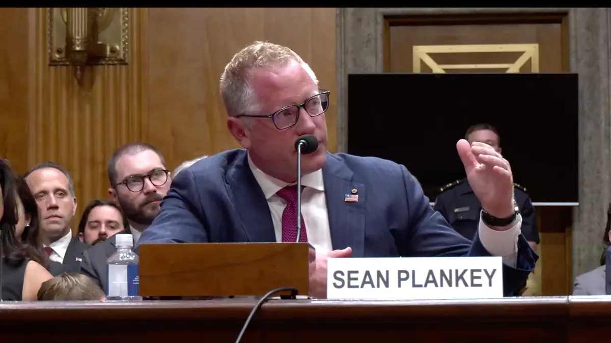 A man gestures as he sits at a table behind a nameplate that says "Sean Plankey"