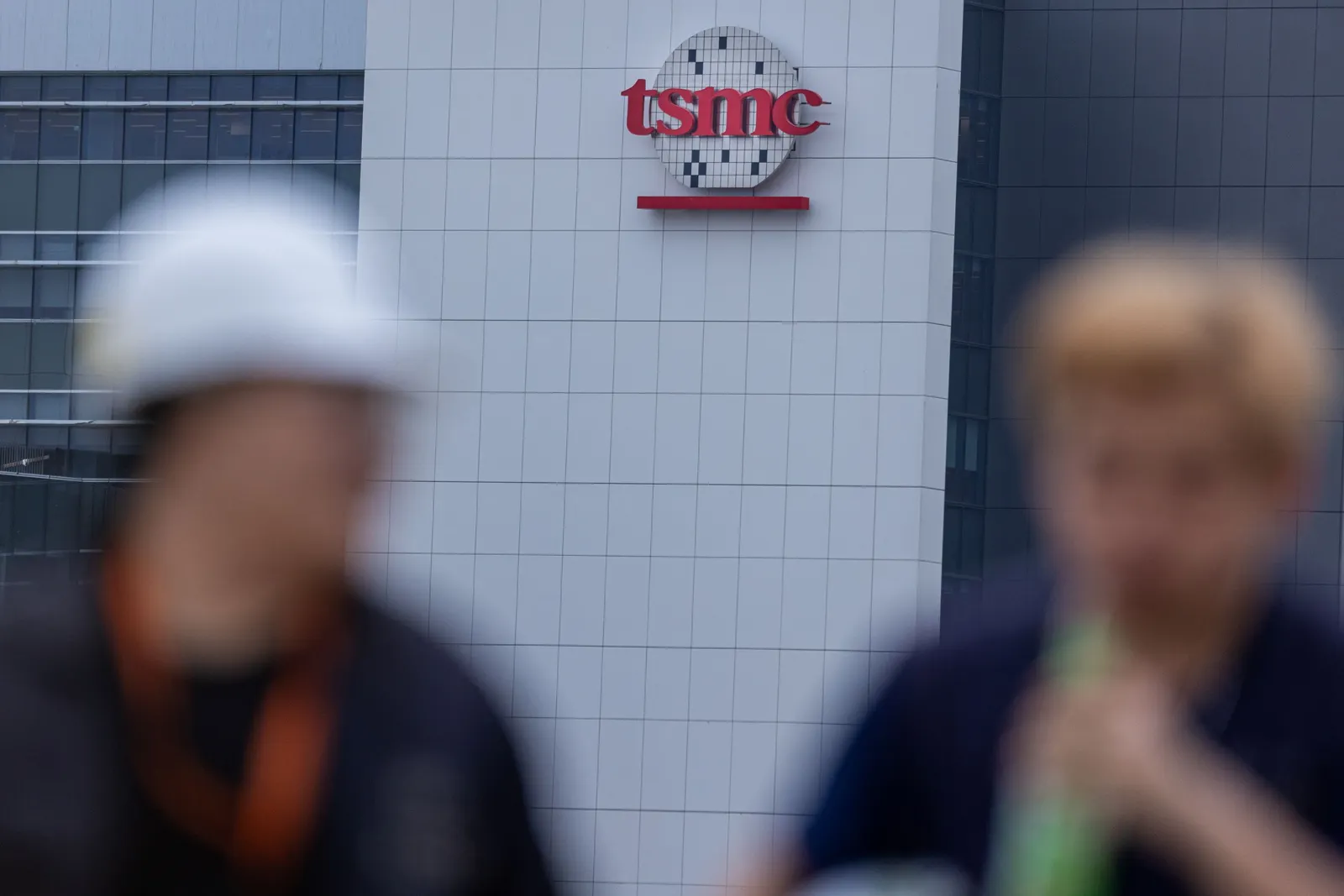 The red, white and black TSMC logo on a building in the background of two blurry individuals.
