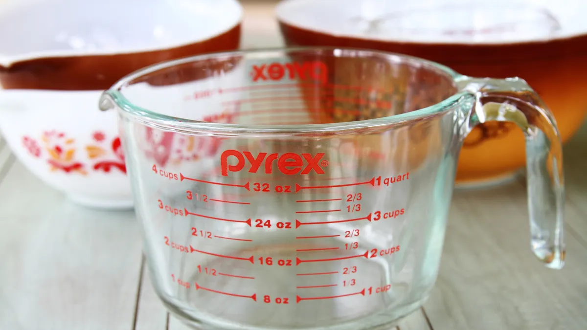 A clear measuring cup with the red Pyrex logo on top of the red measurements in the center, with whit and brown bowls in the background.