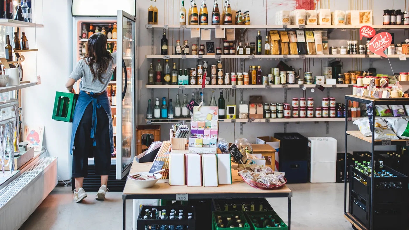 A working stocking a small store, reaching into a fridge while holding a green crate.