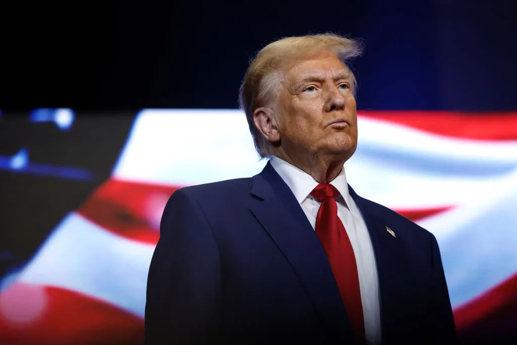 Donald Trump in business attire stands in a stage with an U.S. flag in the background