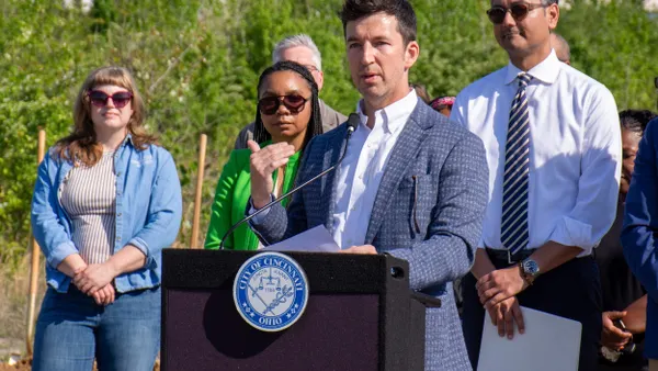 A person wearing a suit speaking at a podium surrounded by other people.