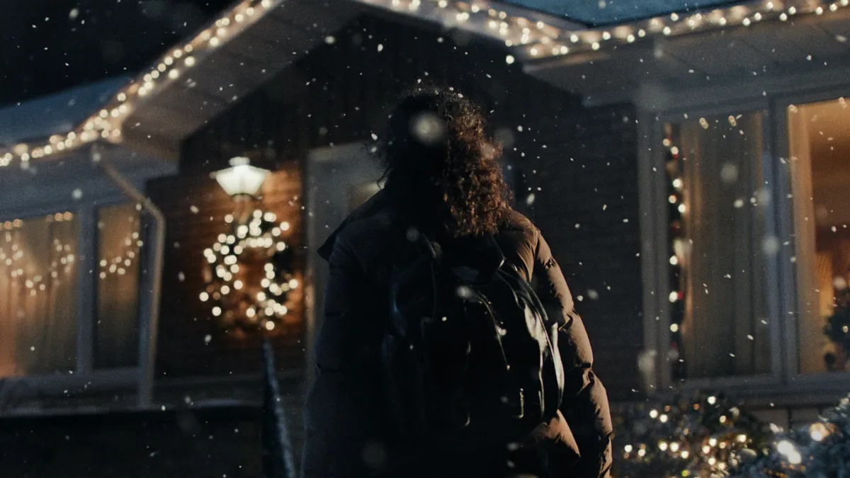 A person wearing a black puffer jacket is shown from the back facing her suburban home