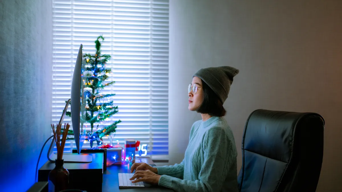 A young person sits at a desk in front of a computer while a Christmas tree is in the background.