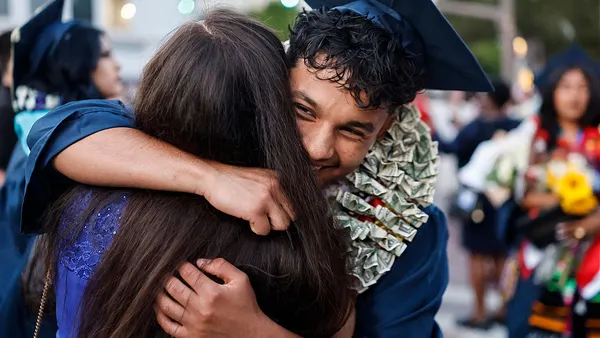 A student in a graduation cap and gown has their face to the camera and is hugging another person. Other graduates can be seen out of focus in the background.