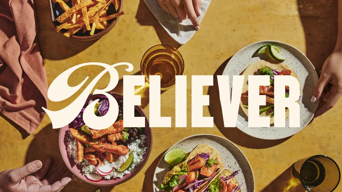 Overhead view of hands touching plates of chicken dishes, including a rice bowl and tacos, on a yellow table with a light brown cloth napkin, two glasses and a bowl of tortilla chips