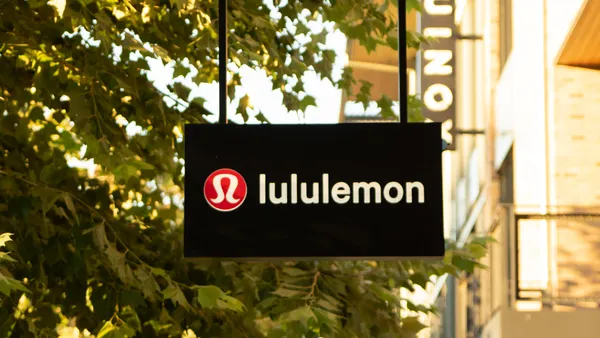 A black "Lululemon" sign features the red and white brand logo and white lettering, hanging in front of a leafy tree.