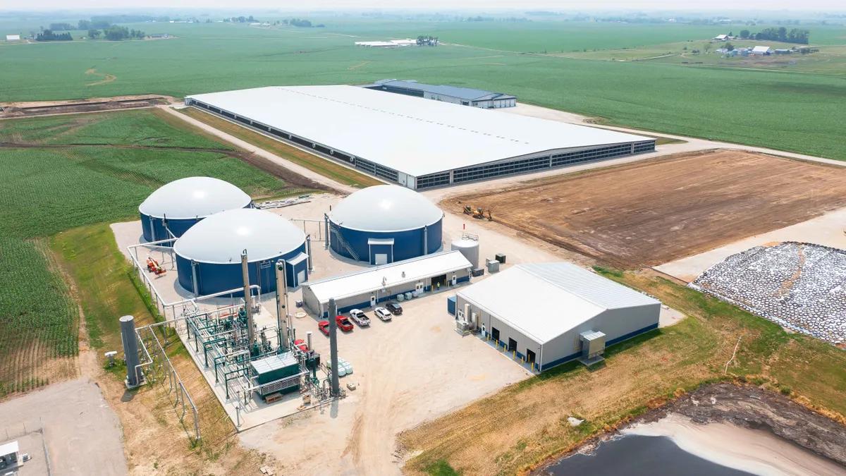 An industrial facility with three circular tank buildings sits near a vast shed and surrounding fields.