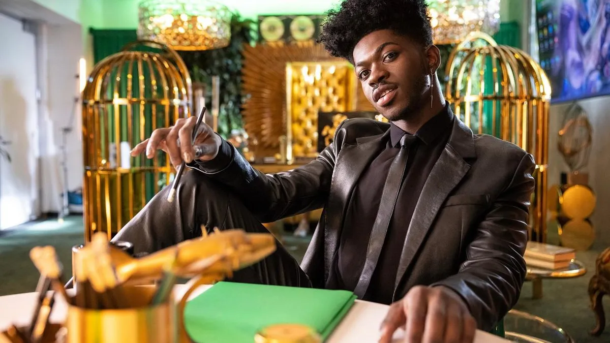 Lil Nas X sits in a gold room wearing a black suit.