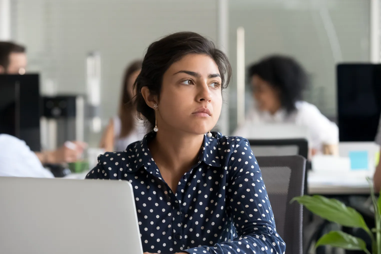 An office worker in a polka-dotted shirt sits at her computer and looks thoughtfully in the distance.