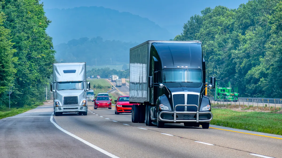 Trucks and passenger cars on an interstate with green trees in the background.