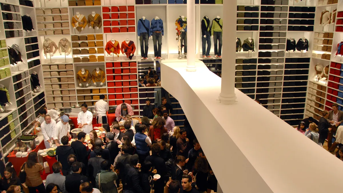 Picture shows the interior of a large two story atrium store as seen from above, filled with clothes on white shelves and crowded with people.