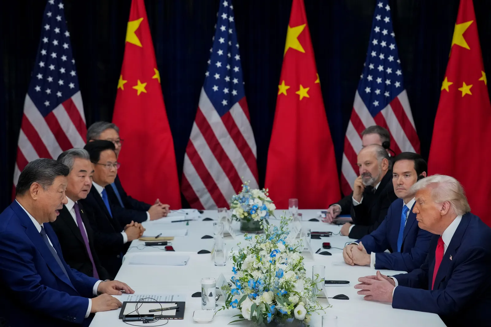 Eight people sit at a table with blue and white flowers and six flags in the background.