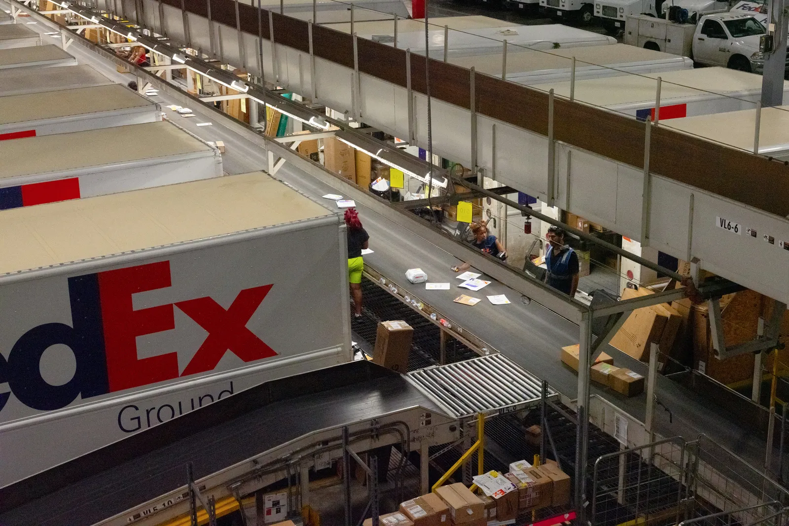 An overhead view of a FedEx warehouse where employees sort packages from a moving conveyor belt and load them into a line of FedEx trucks. The "FedEx Ground" logo is visible on the side of a truck in the foreground.
