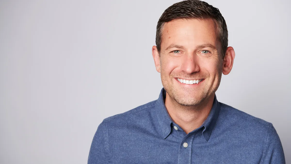 A smiling person with short hair, a light beard and a blue shirt.