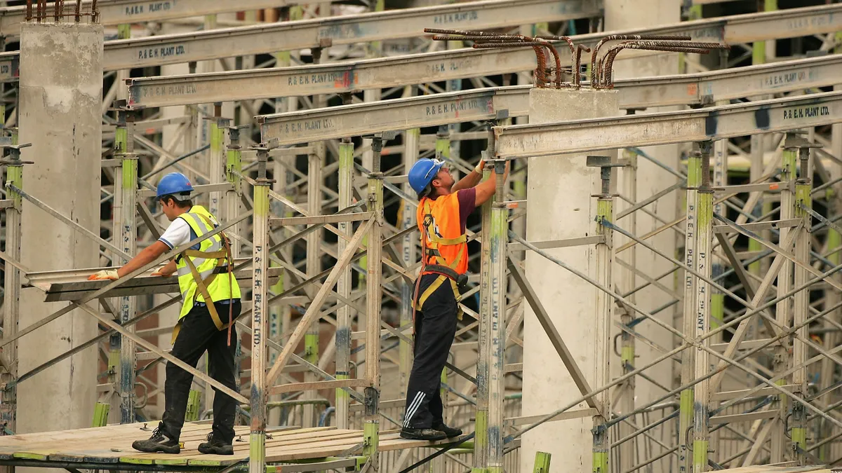 Two construction workers in reflective vests and blue hard hats stand on scaffolding.