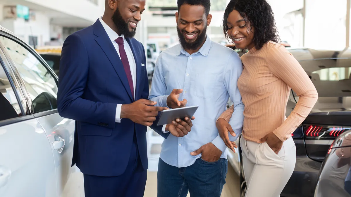 A smartly-dressed male auto sales representative is showing a digital tablet to a young Black couple in a car showroom.