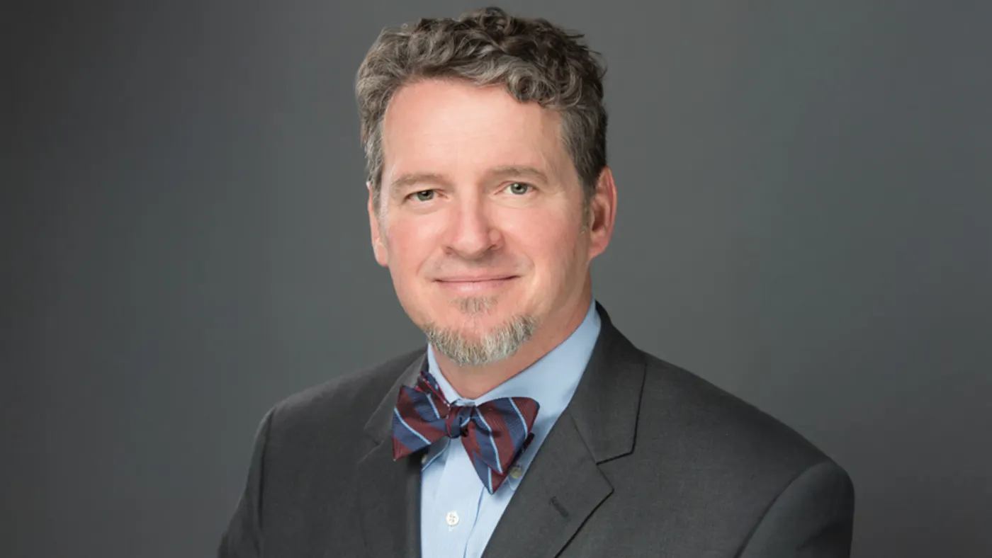 HCA Healthcare's Dr. Thomas Payne wears a gray suit jacket and blue and burgundy striped bow tie, against a gray background.