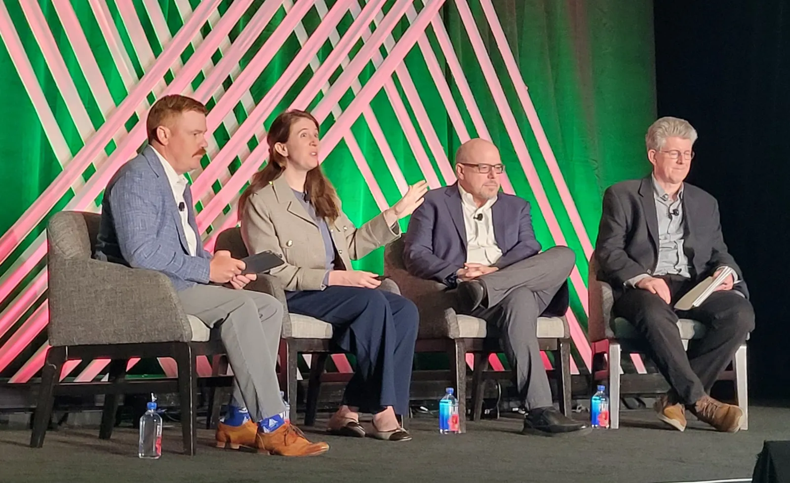 Four people sit in chairs on a stage while speaking to an audience.