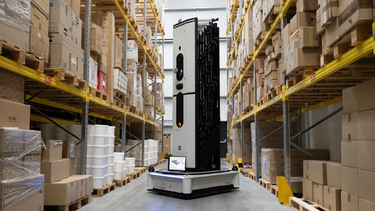 A tall autonomous robot navigates a warehouse aisle lined with shelves of boxes and goods.