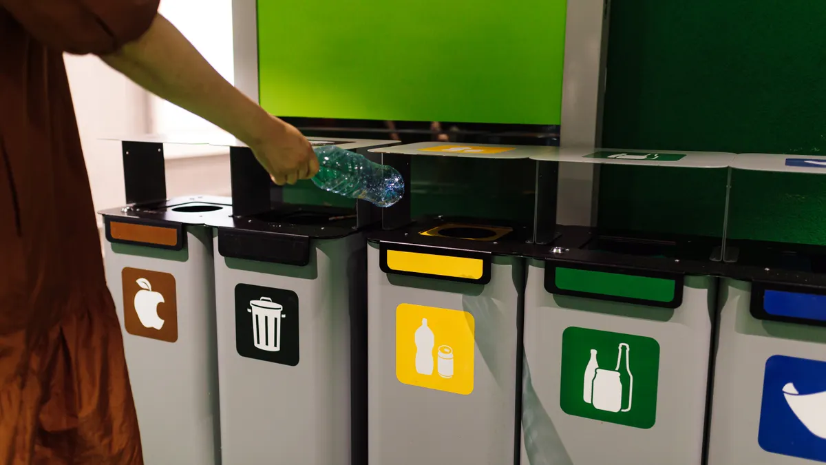 A person's arm is seen throwing a plastic bottle into an appropriate recycling bin, which is next to other waste bins.