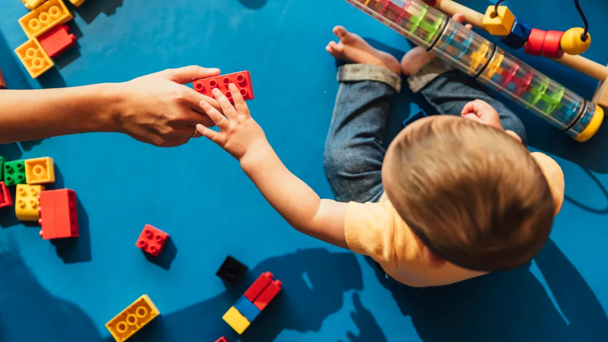 A camera looks down on baby sitting on blue mat and reaching for a colorful block