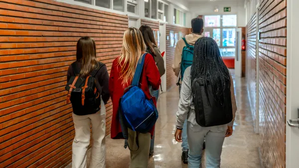 From behind, five students carrying backpacks walk down a school hallway.