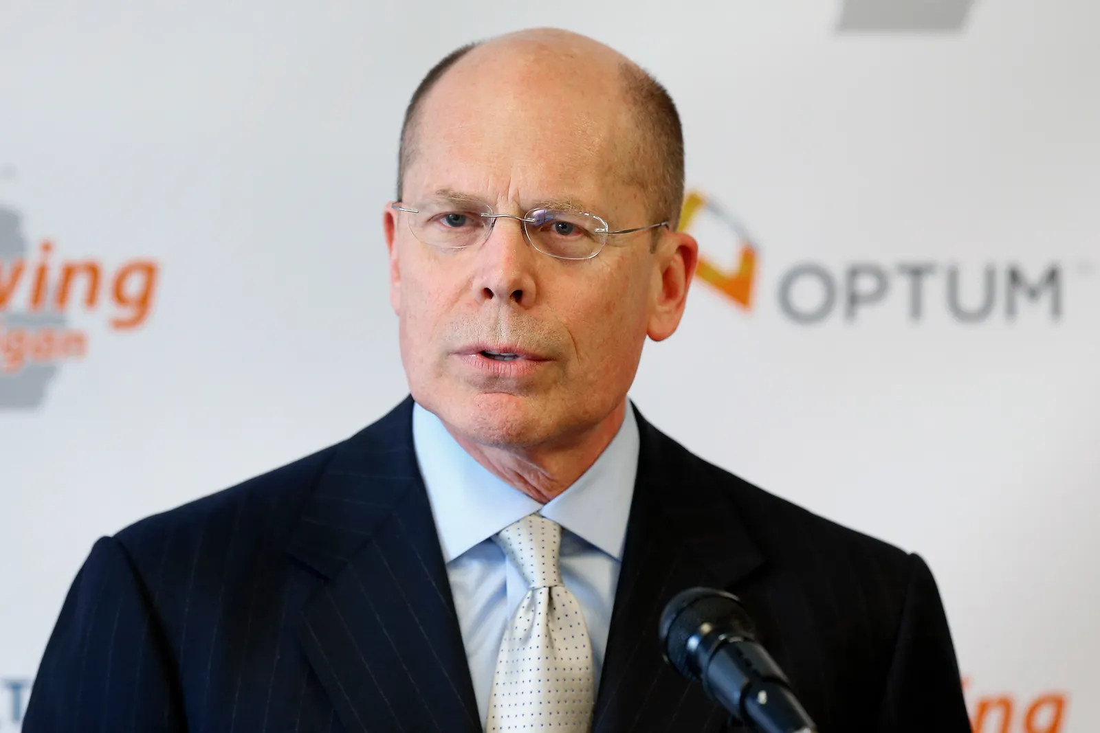 A man in business attire speaks into a microphone, with a backdrop reading “Optum.”