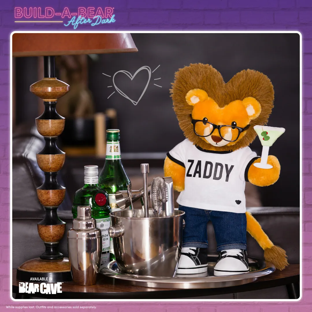 A lion plush holding a martini glass and wearing a &quot;Zaddy&quot; shirt next to a bar set.