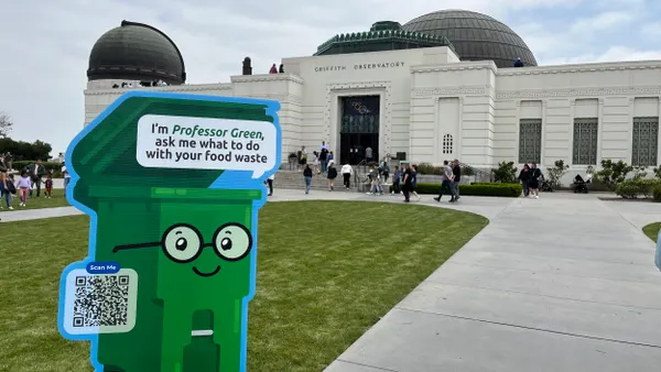 A cardboat cutout of a green cart wearing glasses that says “I’m Professor Green, ask me what to do with your food waste" in front of Griffith Observatory.