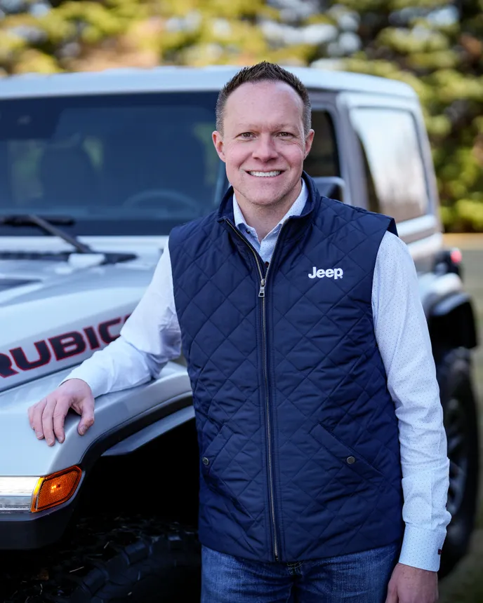 A man in a vest stands near a Jeep