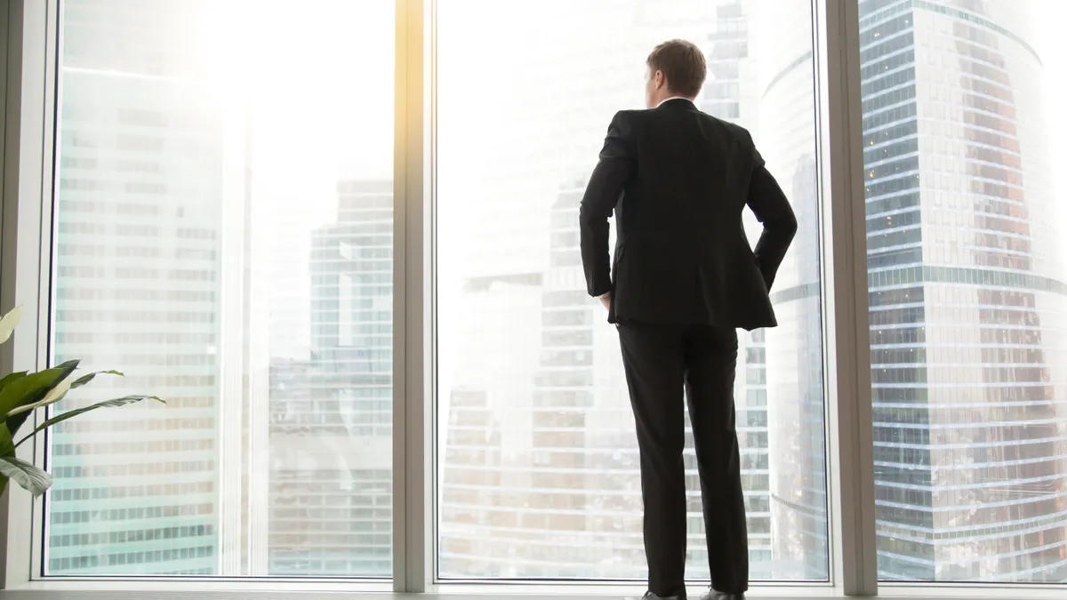 Full length back view of successful businessman in suit standing in office with hands on his waist, CEO looking through window at big city buildings, planning new project, waiting for meeting to start