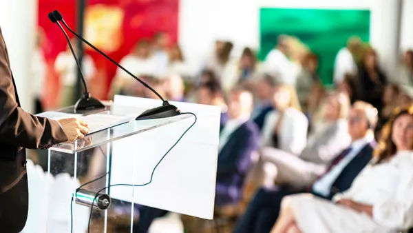 a shoulder height view of a business-clad speaker on a podium in front of an audience at a conference