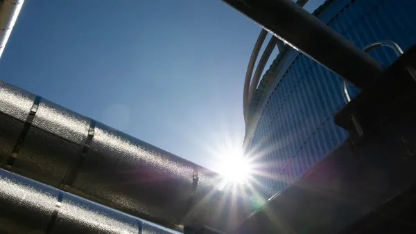 Sunlight shining between large industrial metal pipes and a blue corrugated structure, creating lens flare against a clear sky.