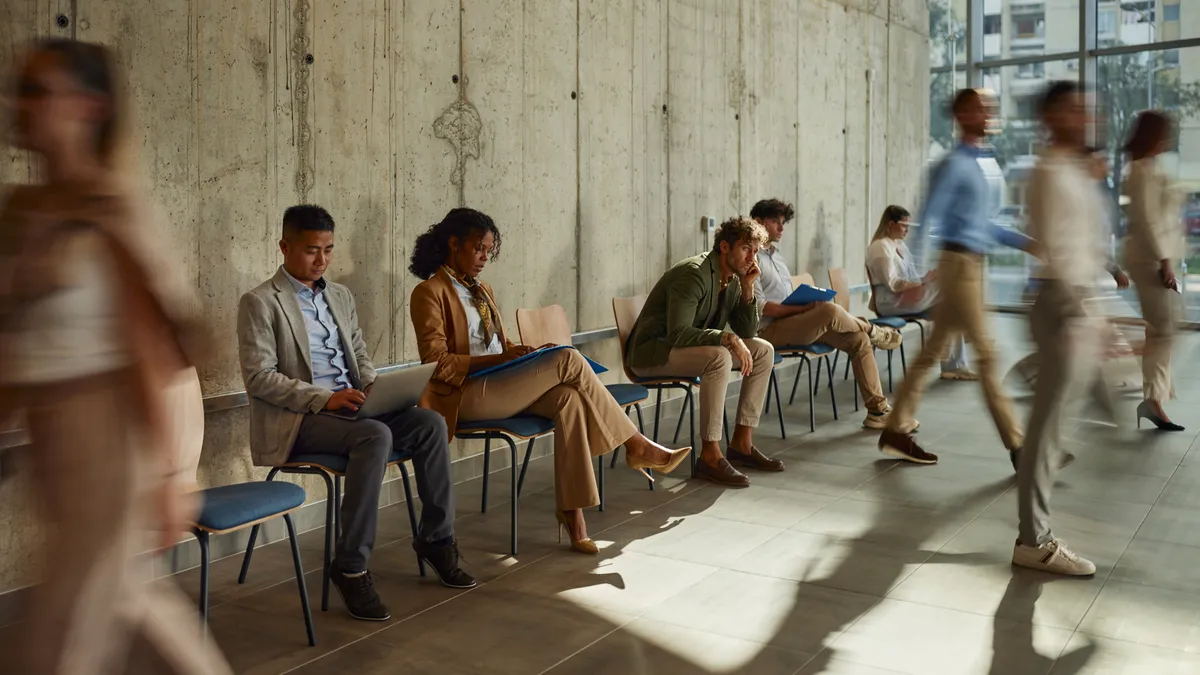 People sit in chairs against a wall while some get up to walk.