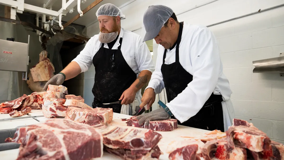 Two men in aprons and hairnets cut slabs of meat