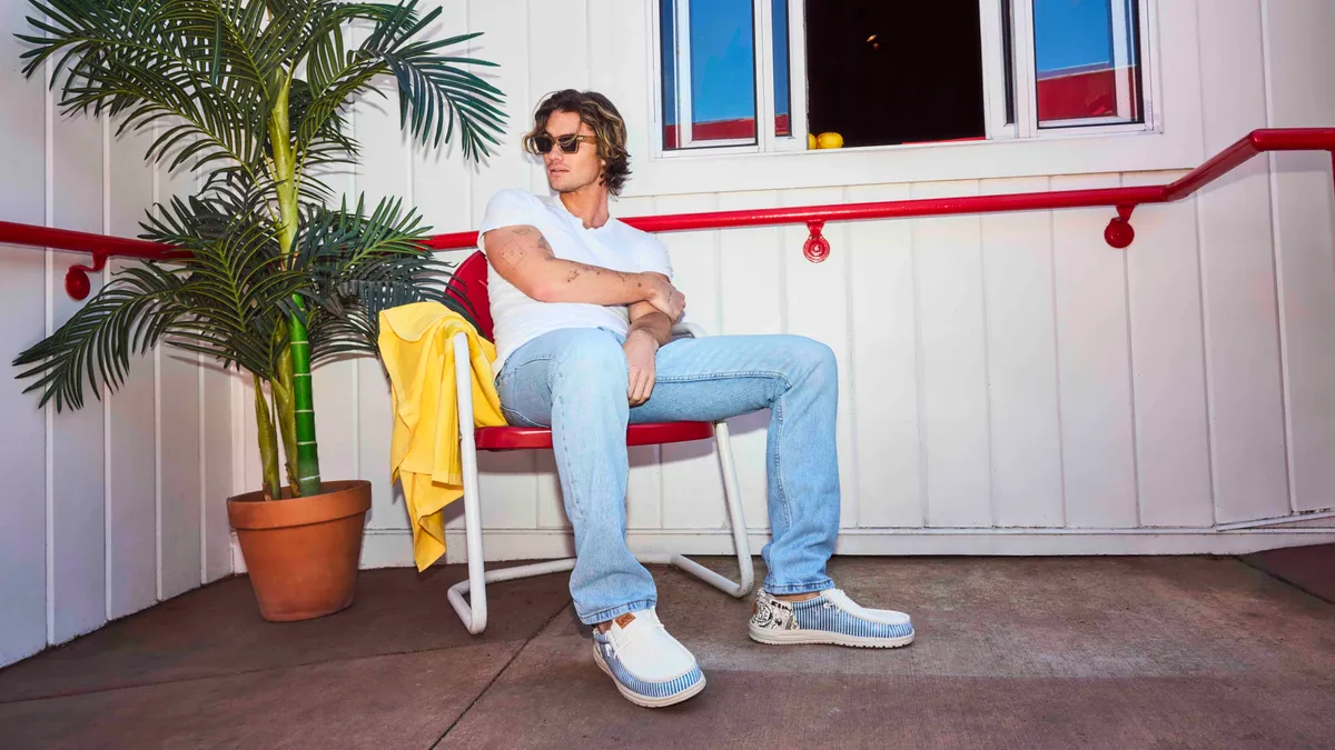 A person wearing a white T-shirt, jeans and sunglasses sits in a red chair against a white wall staring a potted palm tree.