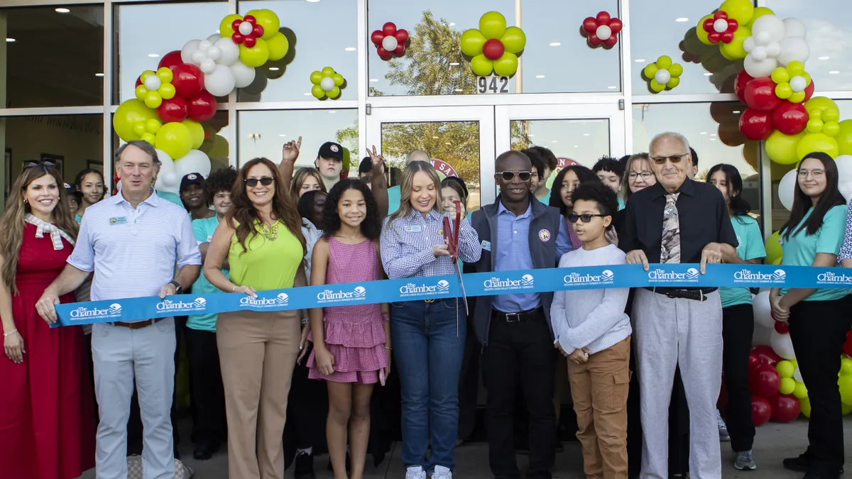 A photograph of a group of people standing behind a ribbon. A person in the middle holds big scissors that are about to cut the ribbon.