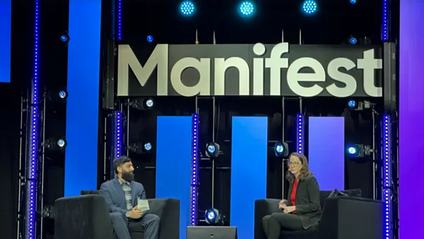 Two panelists sit in chairs while a sign reading "Manifest" hangs above them in the background.