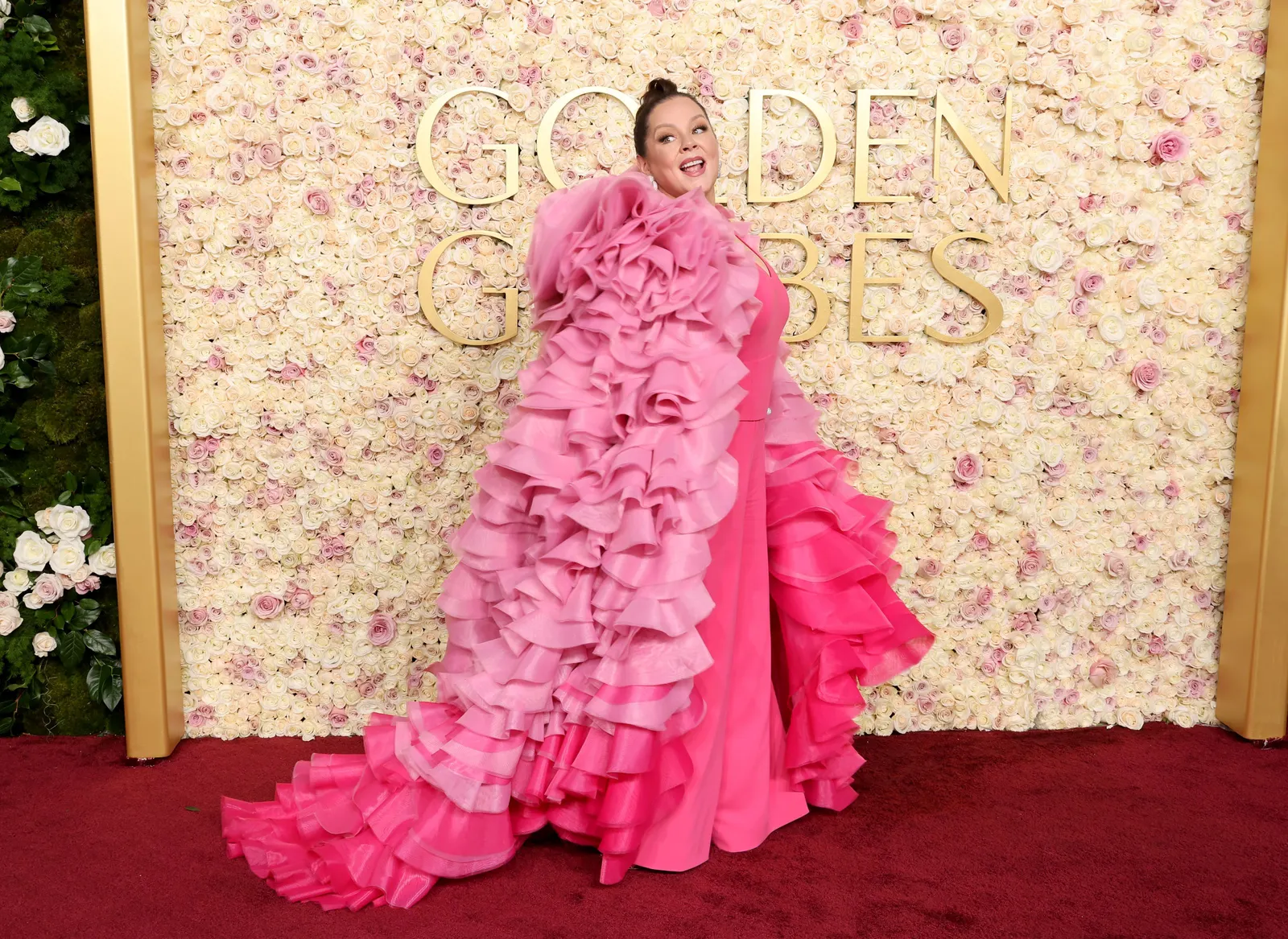 A person in a pink ruffled jacket stands in front of a floral sign that reads Golden Globes.