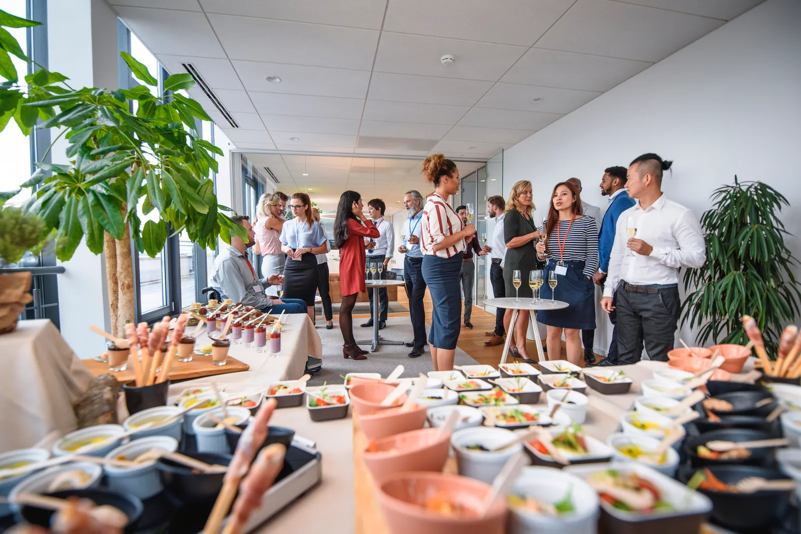 Small dishes of savory and sweet foods on buffet tables and business professionals in background enjoying sparkling wine at office party.